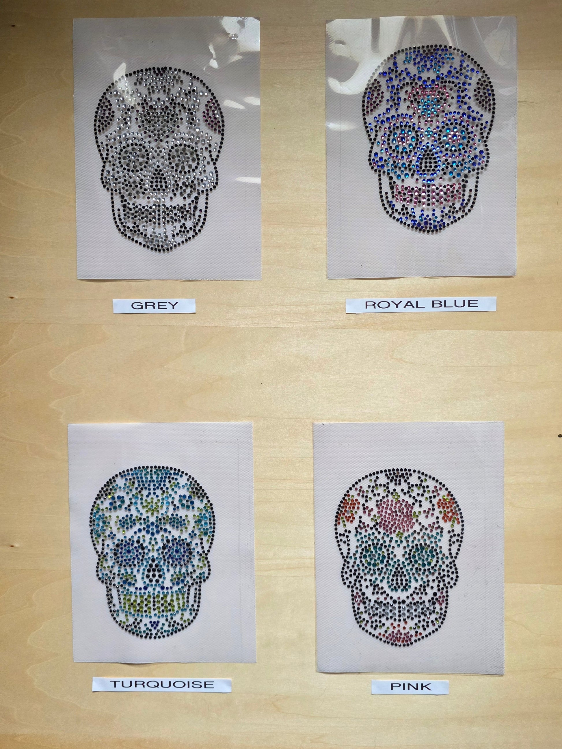 A wooden display board showing four skull designs labelled Grey, Royal Blue, Turquoise, and Pink, each made of small colourful rhinestones, ready to iron on to any garment.