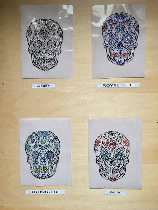 A wooden display board showing four skull designs labelled Grey, Royal Blue, Turquoise, and Pink, each made of small colourful rhinestones, ready to iron on to any garment.
