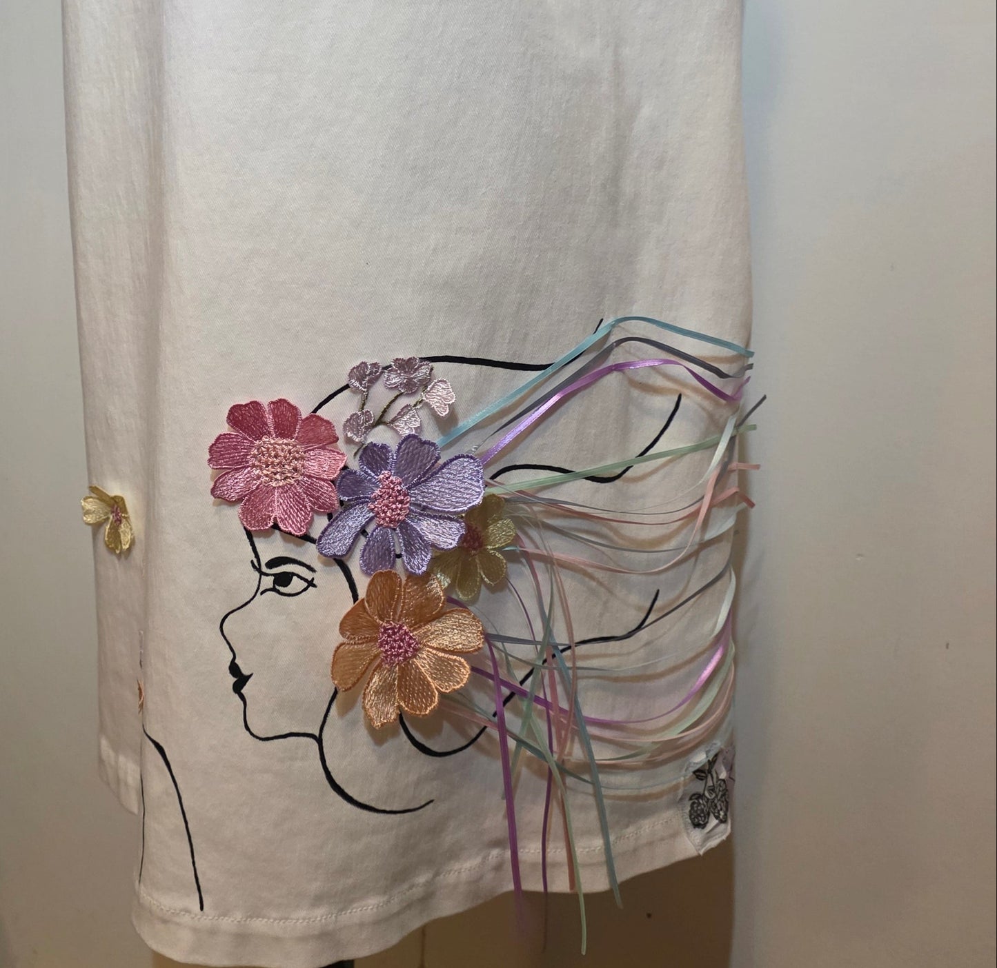 White denim sleeveless dress hand embellished with line-art woman, dimensional embroidered flowers and cascading pastel satin ribbons.